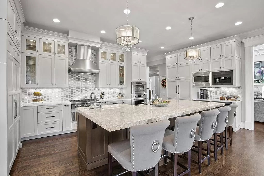Cumming, GA area kitchen remodel with white shaker cabinets to the ceiling, glass-front uppers, a large stone-topped island seating five, twin drum chandeliers, a stainless range and mosaic tile backsplash